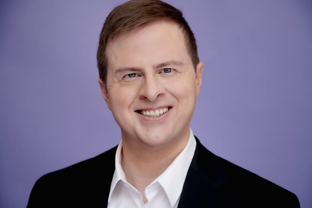 Portrait of Stephen Letnes wearing a dark suit with white shirt, with a purple background